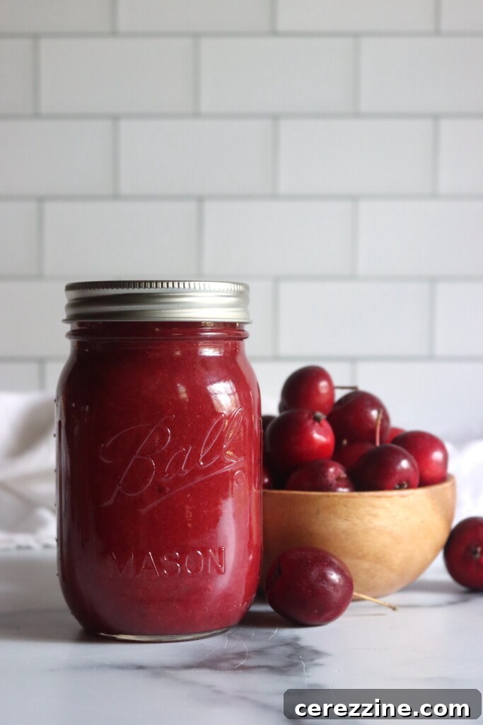 A jar of home canned crabapple sauce on a white table next to a bowl of bright red dolgo crabapples.