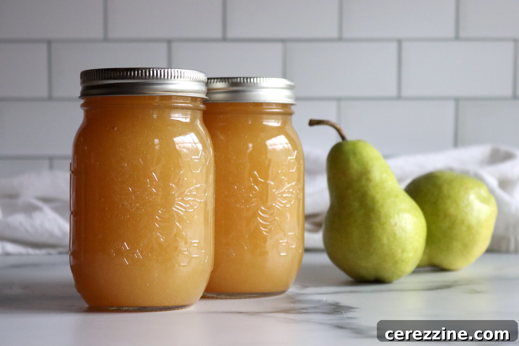 Two jars of pear honey with fresh green pears on a marble counter