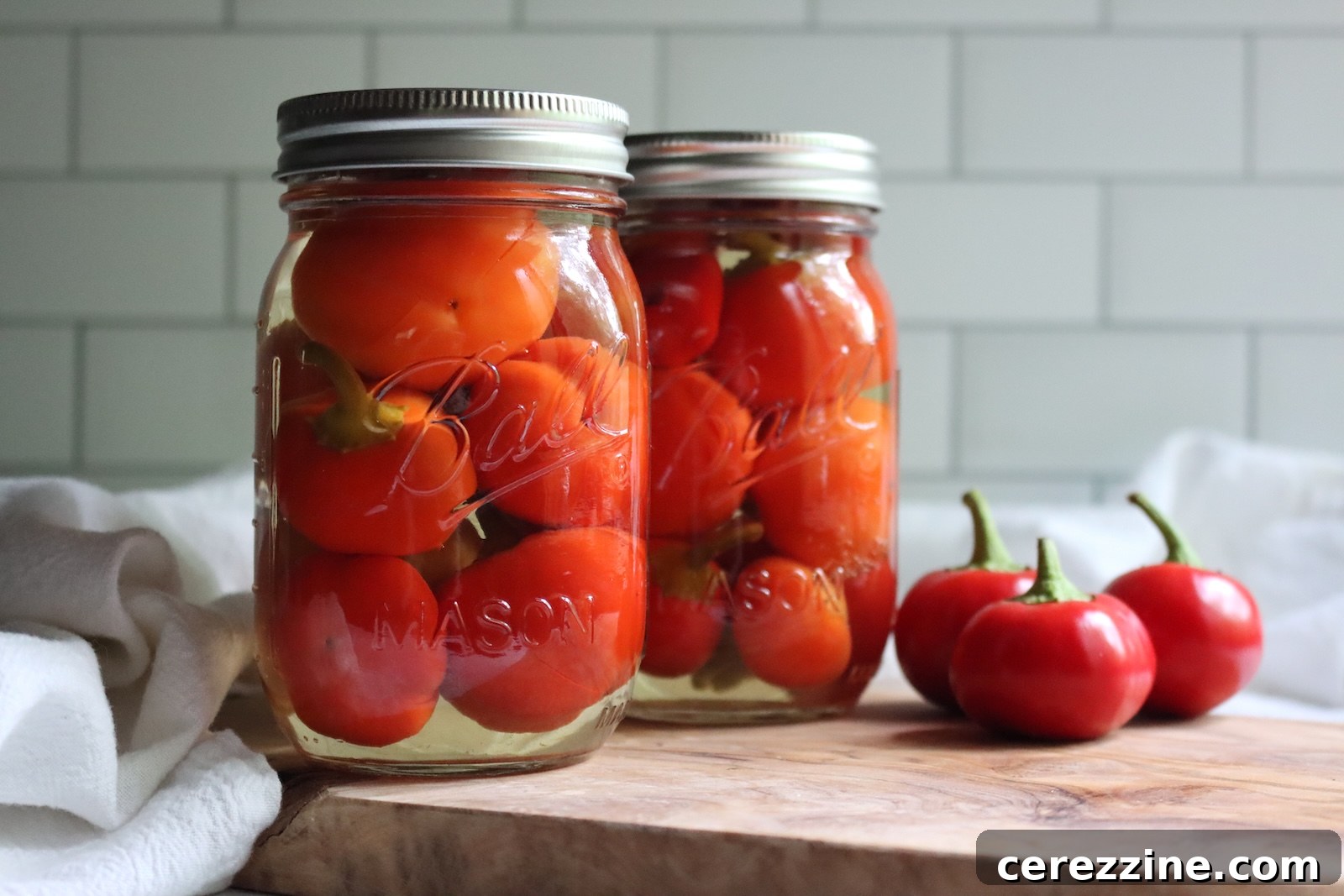 Two jars of pickled cherry peppers on a wooden board with fresh cherry peppers beside them