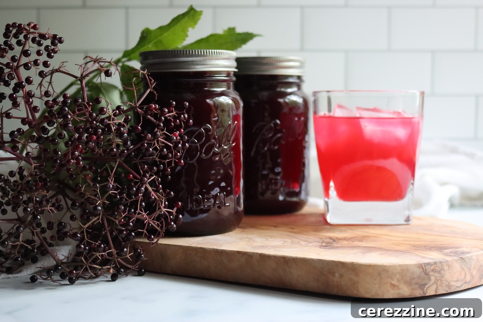 Two jars of elderberry lemonade concentrate and a glass of elderberry lemonade over ice on a wooden board