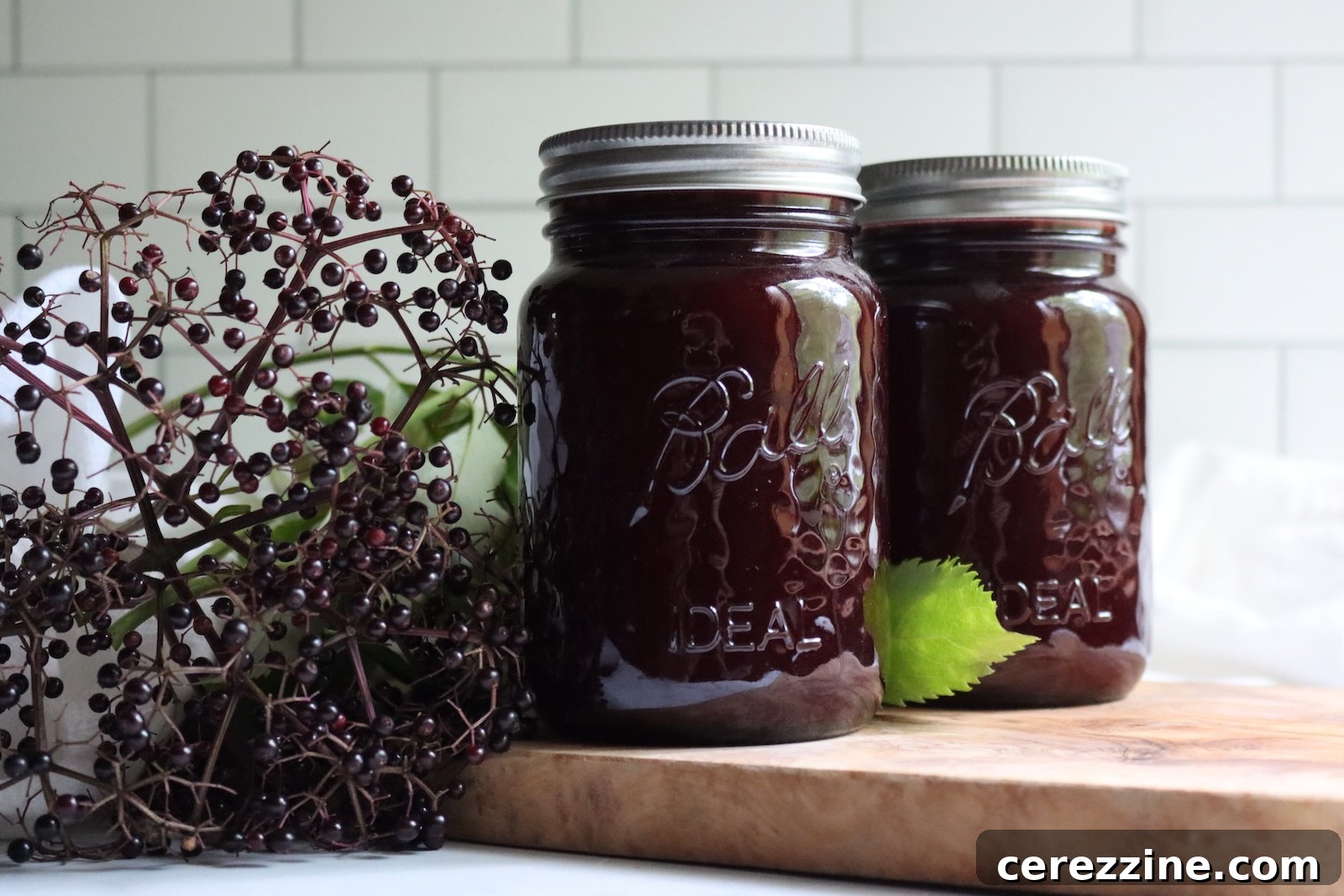 Two sealed jars of canned elderberry lemonade concentrate on a wooden board beside elderberry clusters