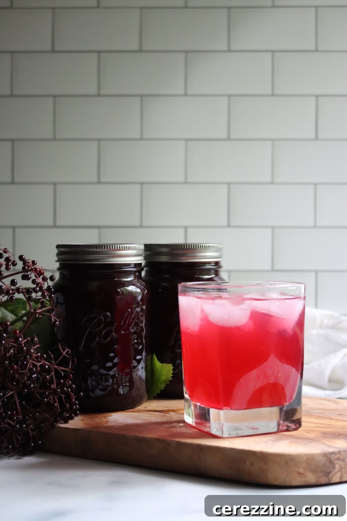Iced glass of pink elderberry lemonade in front of two jars of concentrate on a wooden board