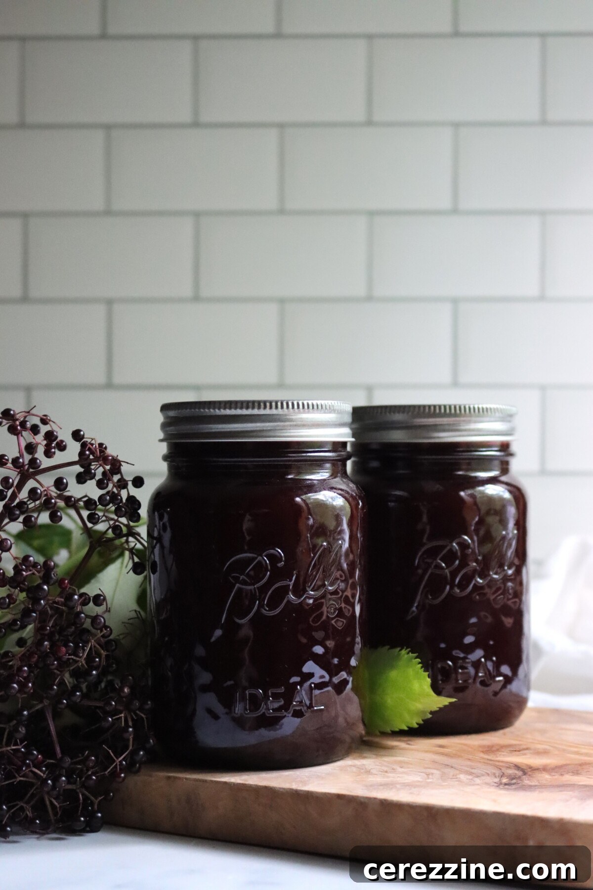 Two jars of homemade elderberry lemonade concentrate on a wooden board next to fresh elderberry clusters