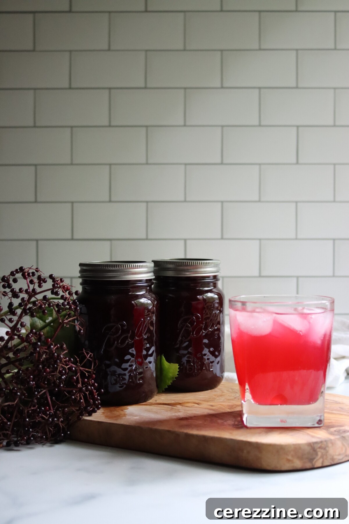 Glass of elderberry lemonade over ice beside two jars of elderberry lemonade concentrate