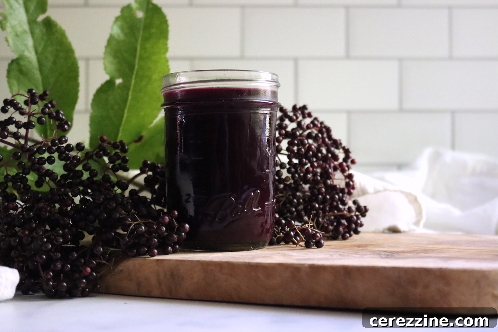 Jar of strained elderberry juice on a wooden board surrounded by fresh elderberry clusters