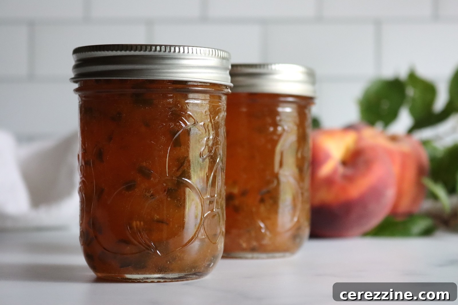 Jars of homemade peach jalapeño jam on a marble counter with ripe peaches