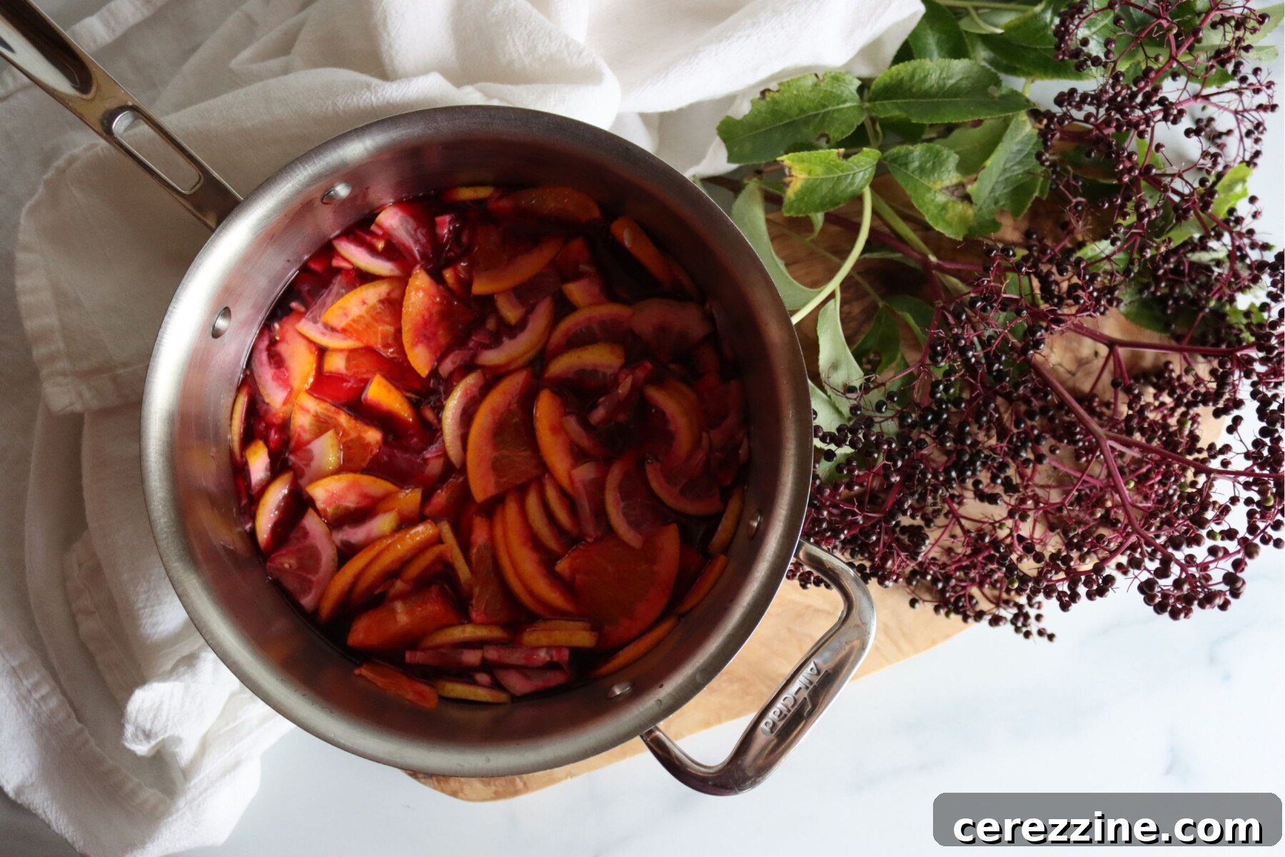 Lemon and orange slices simmering in elderberry juice in a saucepan, with fresh elderberries beside it