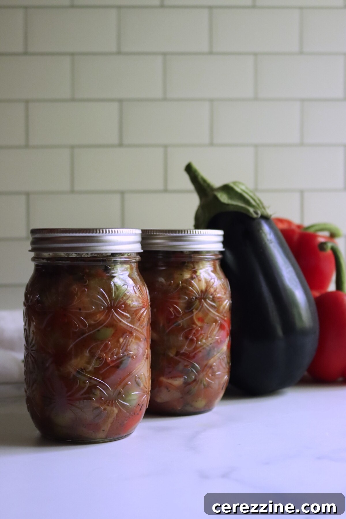 Two pints of eggplant caponata with a fresh eggplant and red peppers in the background