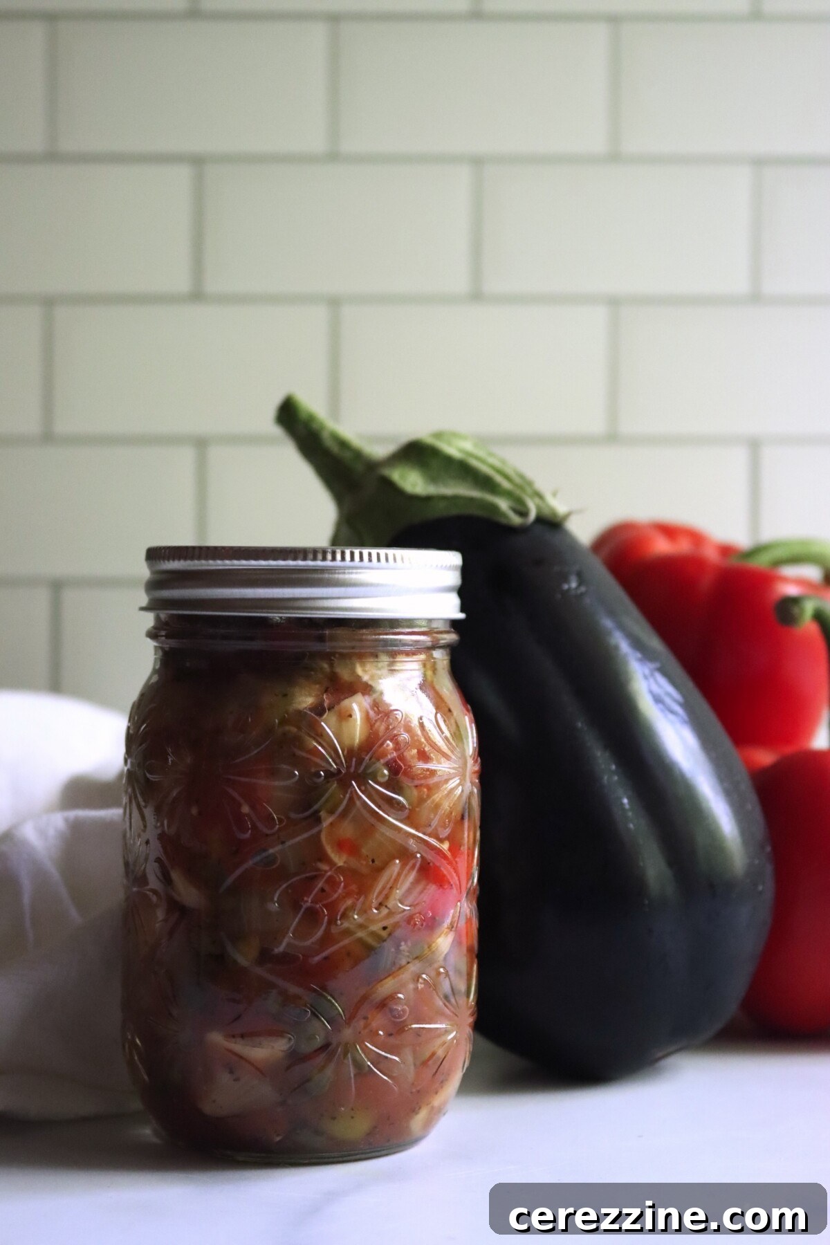 Jar of home-canned eggplant caponata in front of a fresh eggplant and red peppers