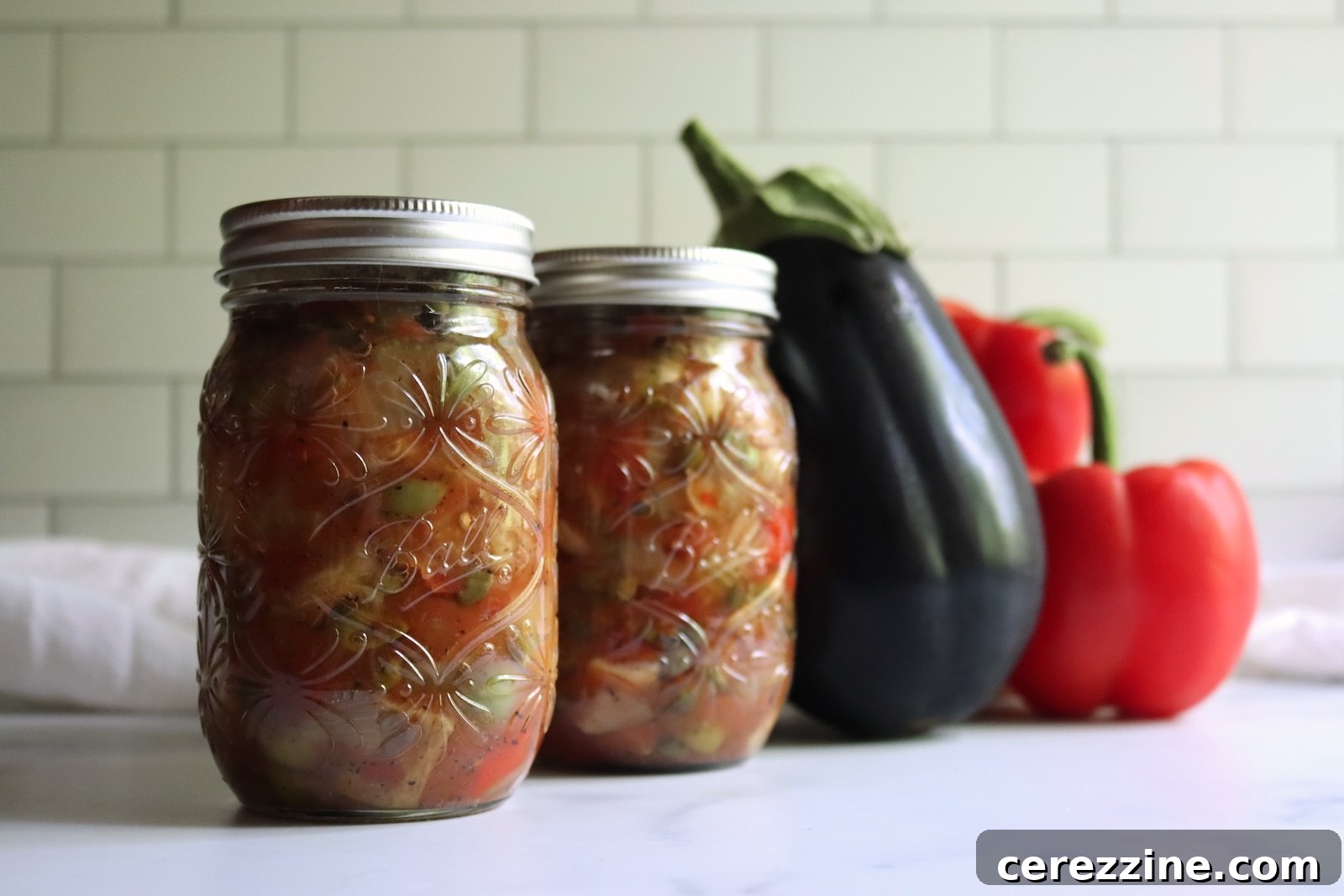Two pint jars of eggplant caponata beside a fresh eggplant and red bell peppers