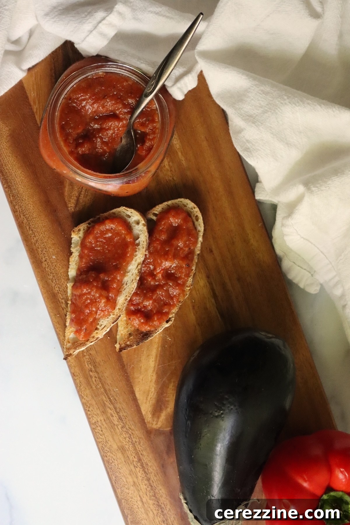 Spoon in an open jar of ajvar next to bread spread with ajvar, an eggplant, and a red pepper