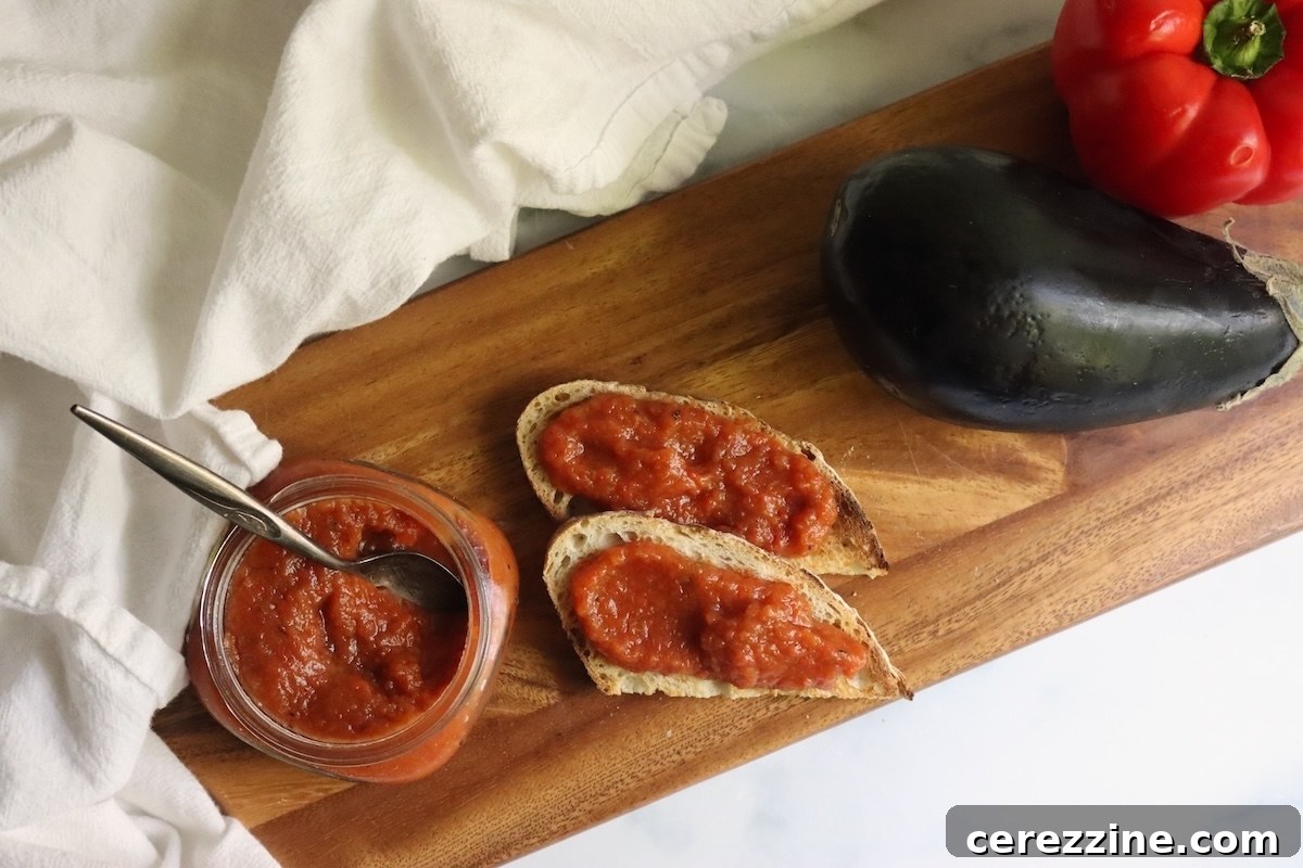 Open jar of ajvar with a spoon beside two slices of bread spread with ajvar on a wooden board