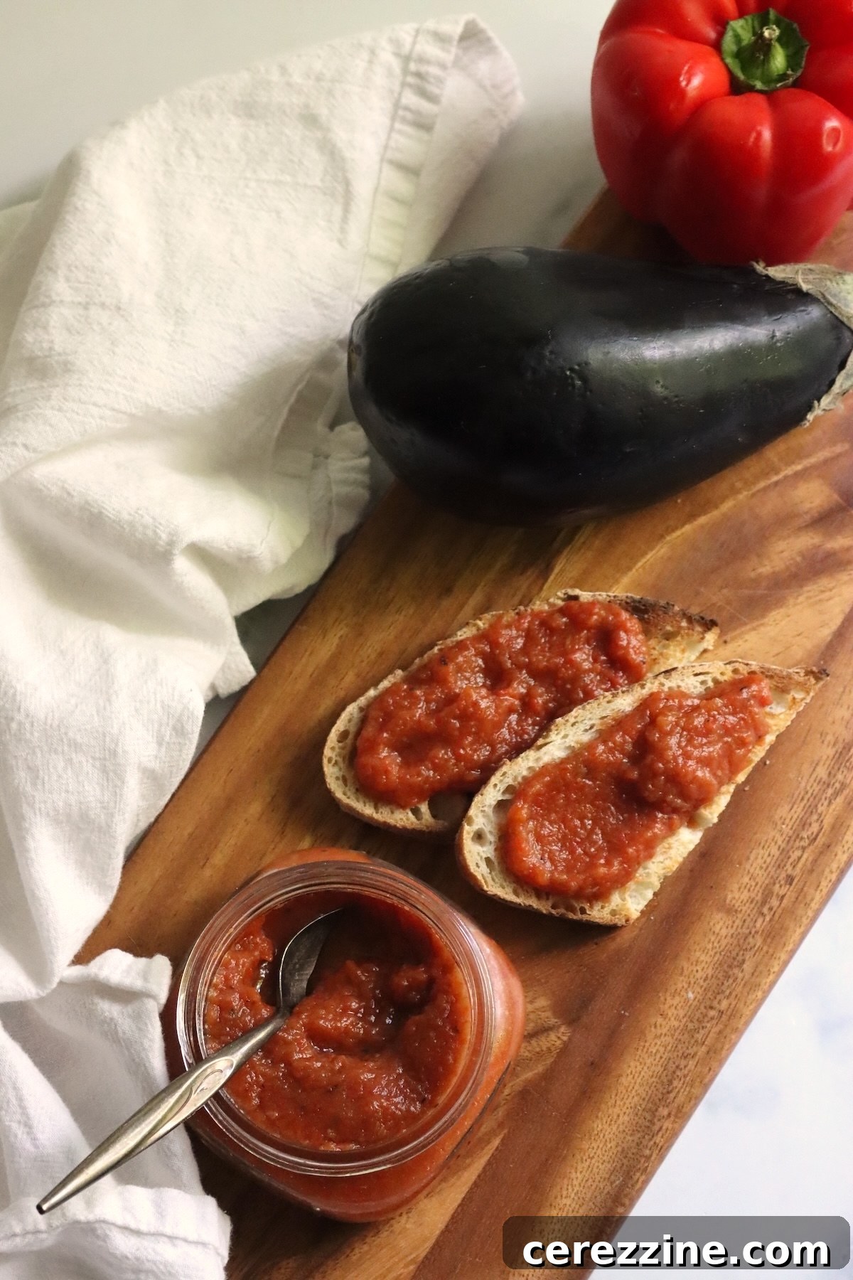 Slices of bread spread with ajvar, an open jar, an eggplant, and a red pepper on a cutting board