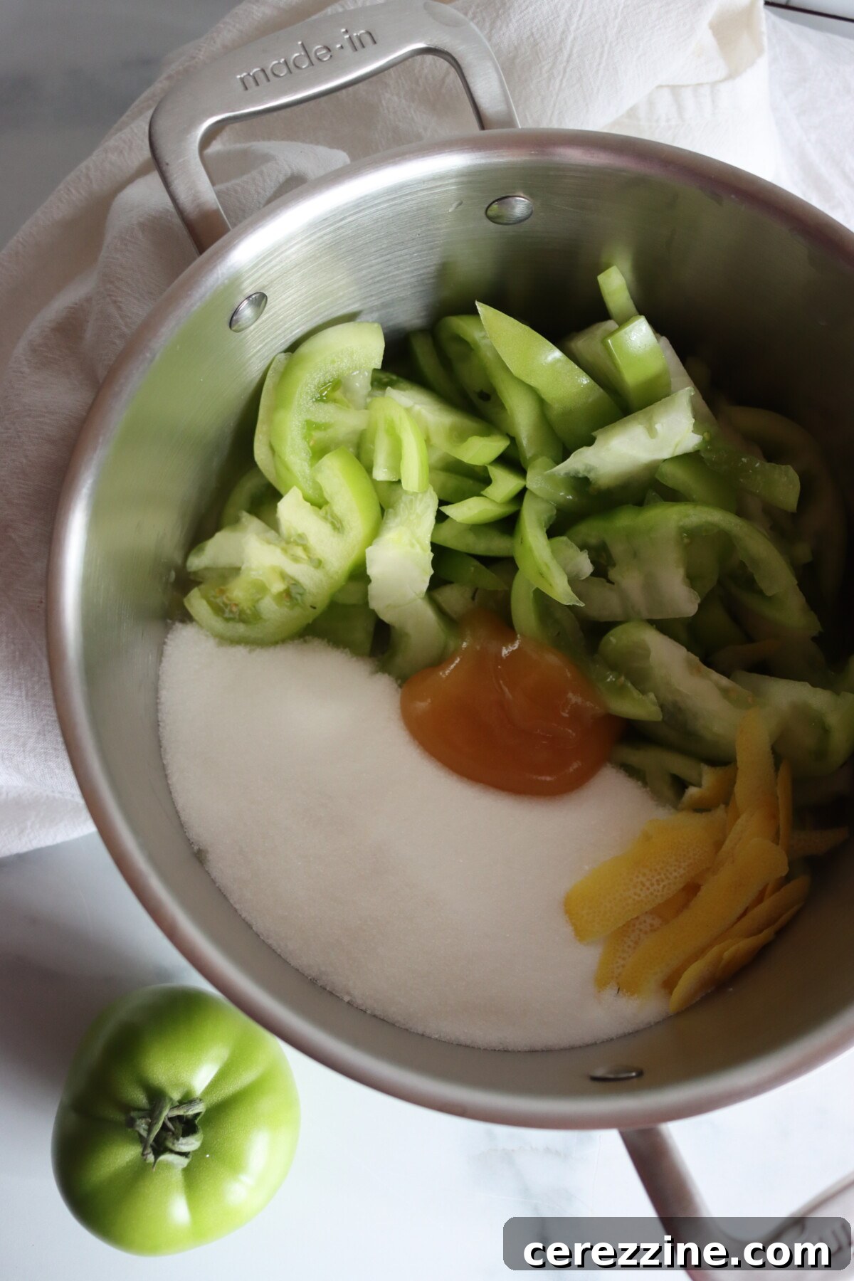 Sliced green tomatoes, sugar, honey, and strips of lemon peel in a stainless steel pot