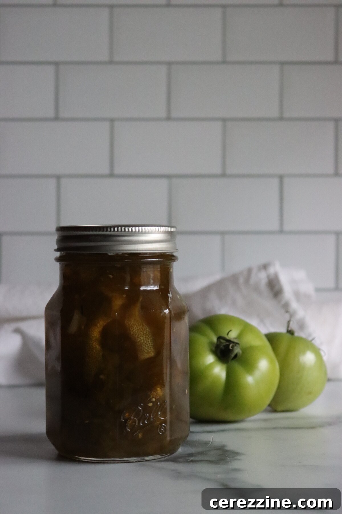 Sealed pint jar of green tomato preserves with green tomatoes behind it