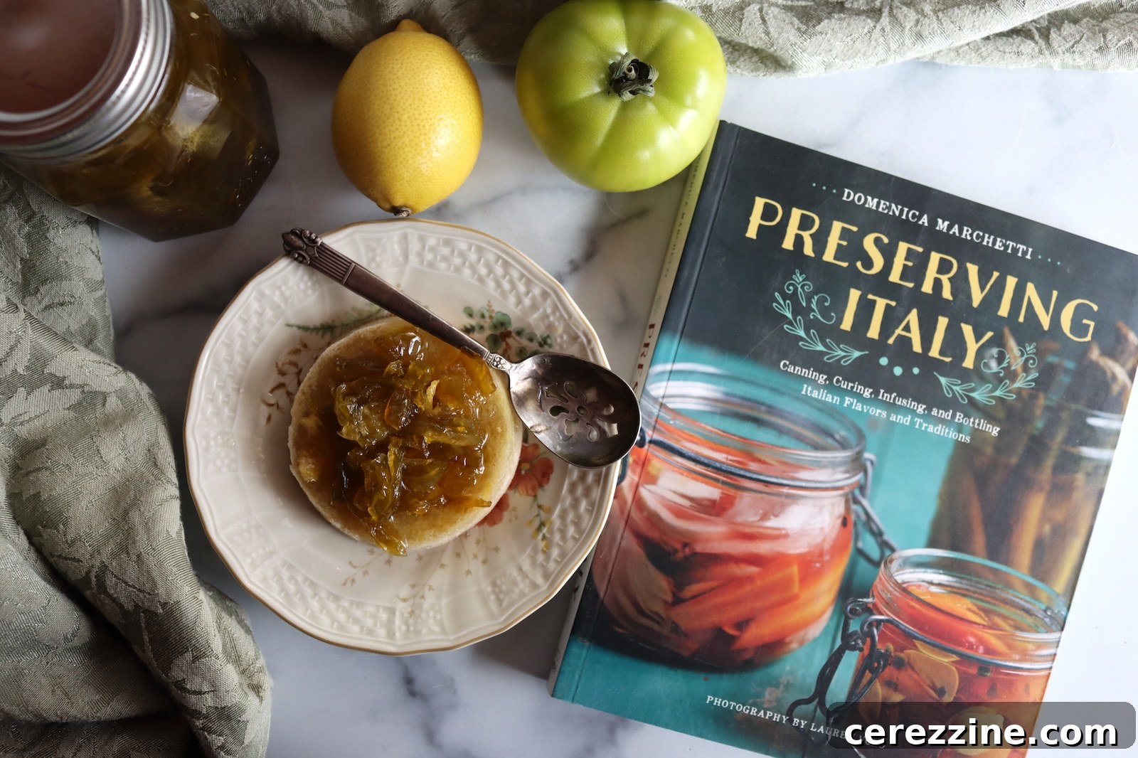 Green tomato preserves on an English muffin with a lemon, a green tomato, and the book Preserving Italy