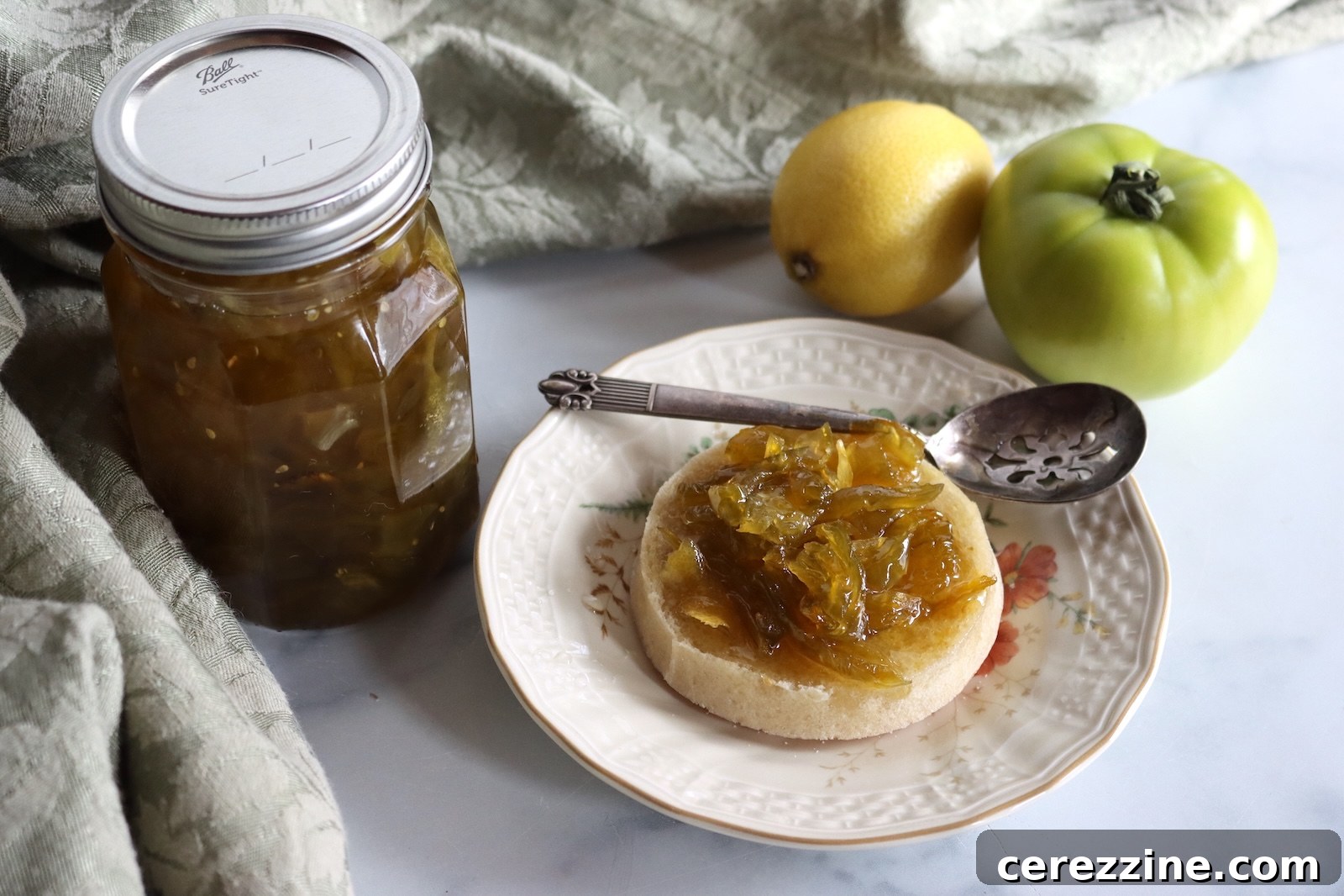 Jar of green tomato preserves with a spoonful on an English muffin, a lemon, and a green tomato